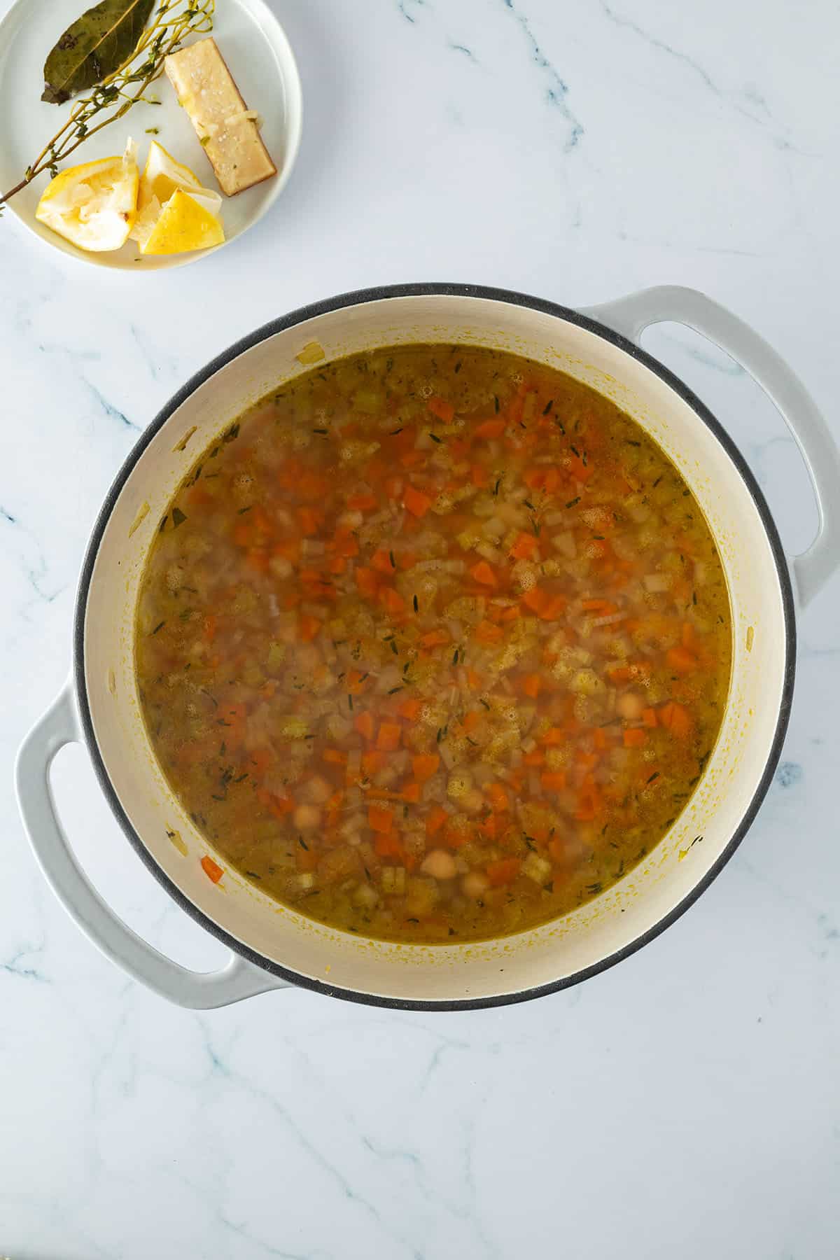 Fertig gekochter Kichererbseneintop in einem Topf, daneben ein kleiner Teller mit einer ausgepressten Zitrone, gekochten Kräutern und einem gekochten Stück Parmesanrinde.