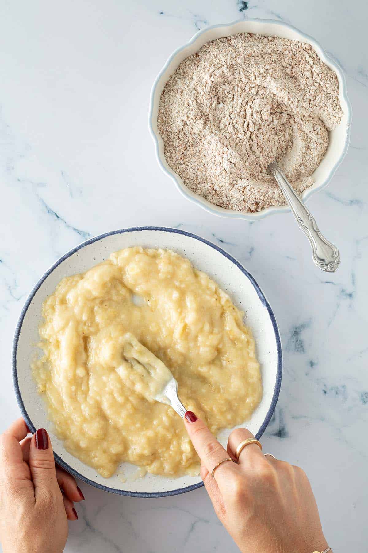 Zwei Schüsseln mit Zutaten für Bananenbrot: eine Schüssel mit zerdrückten Bananen, die mit einer Gabel bearbeitet werden, daneben eine Schüssel mit Mehlmischung und Löffel.