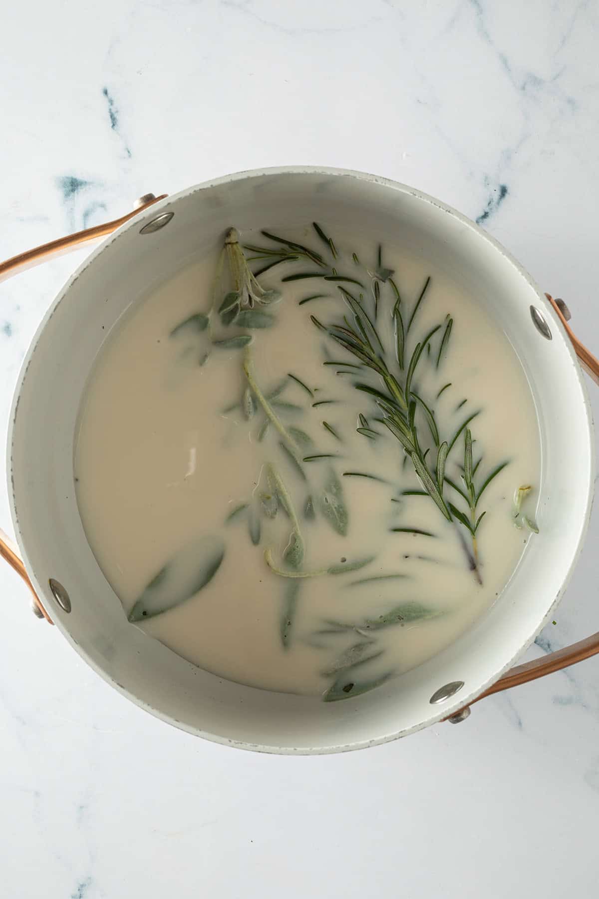 Milch wird mit frischen Rosmarin- und Salbeizweigen in einem Topf erhitzt.