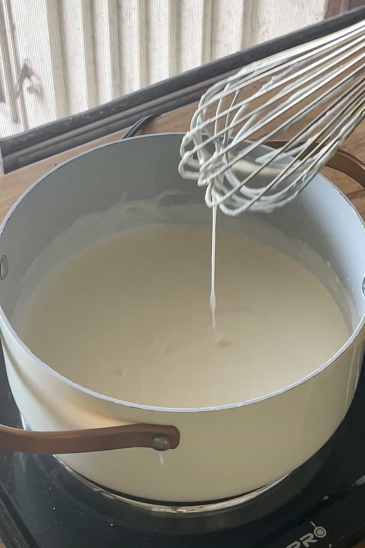 Cremige, weiße Käsesauce in einem Topf, die von einem Schneebesen in einem dünnen Strahl herunterläuft.