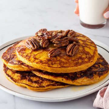 Ein Stapel Kürbis Pancakes auf einem Teller mit Nüssen und Ahornsirup. Eine Hand hält ein Glas Milch im Hintergrund.