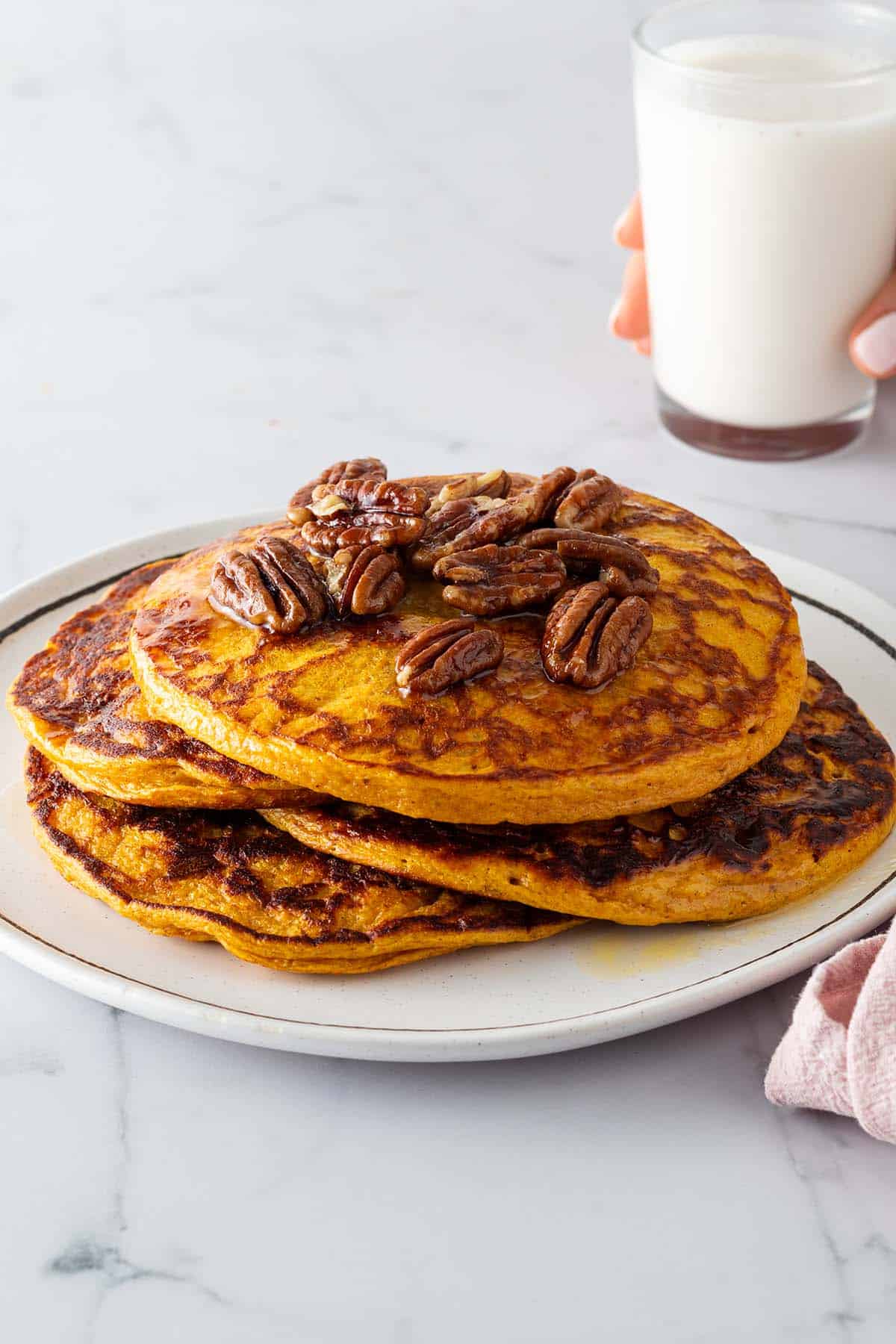 Ein Stapel Kürbis Pancakes auf einem Teller mit Nüssen und Ahornsirup. Eine Hand hält ein Glas Milch im Hintergrund.