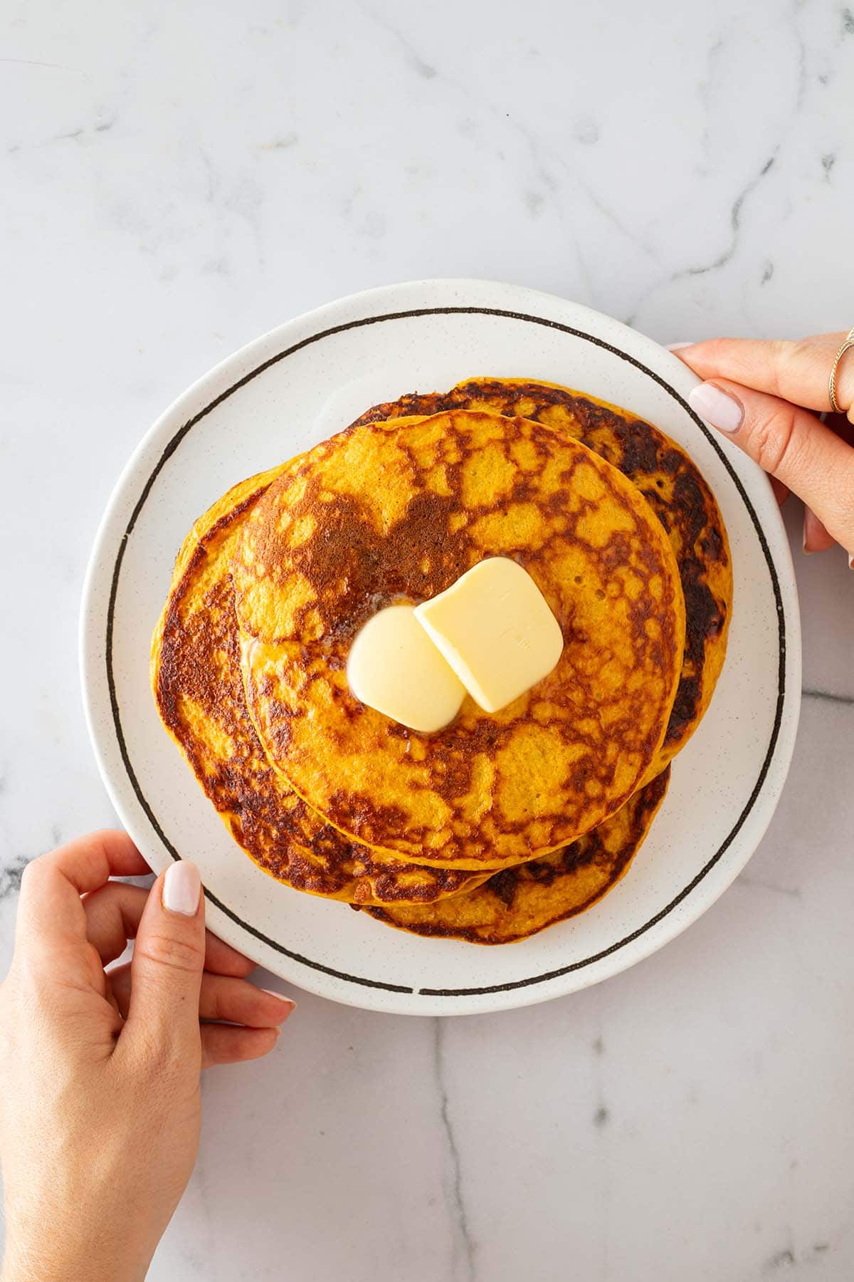 Eine Person hält ein Teller mit mehreren Kürbis Pancakes in der Hand mit zwei Stücke Butter auf dem obersten Pancake.