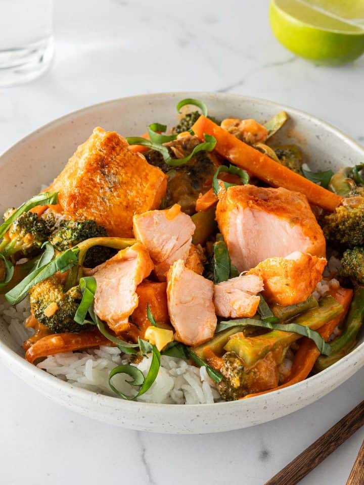 Eine Schüssel mit Thai Lachs Curry mit saftigen Lachswürfeln, Karotten und Brokkoli serviert auf Reis mit frischem Basilikum. Im Hintergrund ein Glas Wasser, eine Limette und Esstäbchen im Bildrand.