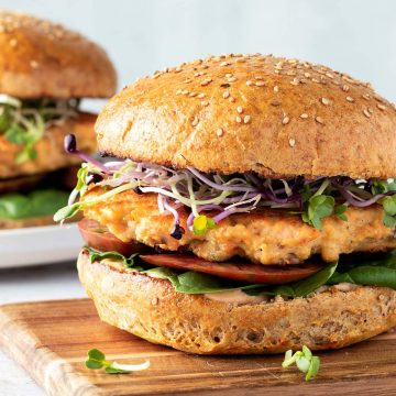 Ein saftiger Lachsburger mit einem goldbraun angebratenen Lachs Patty, frischen Sprossen, Tomaten und Salat auf einem Holzbrett, im Hintergrund ein zweiter Burger.