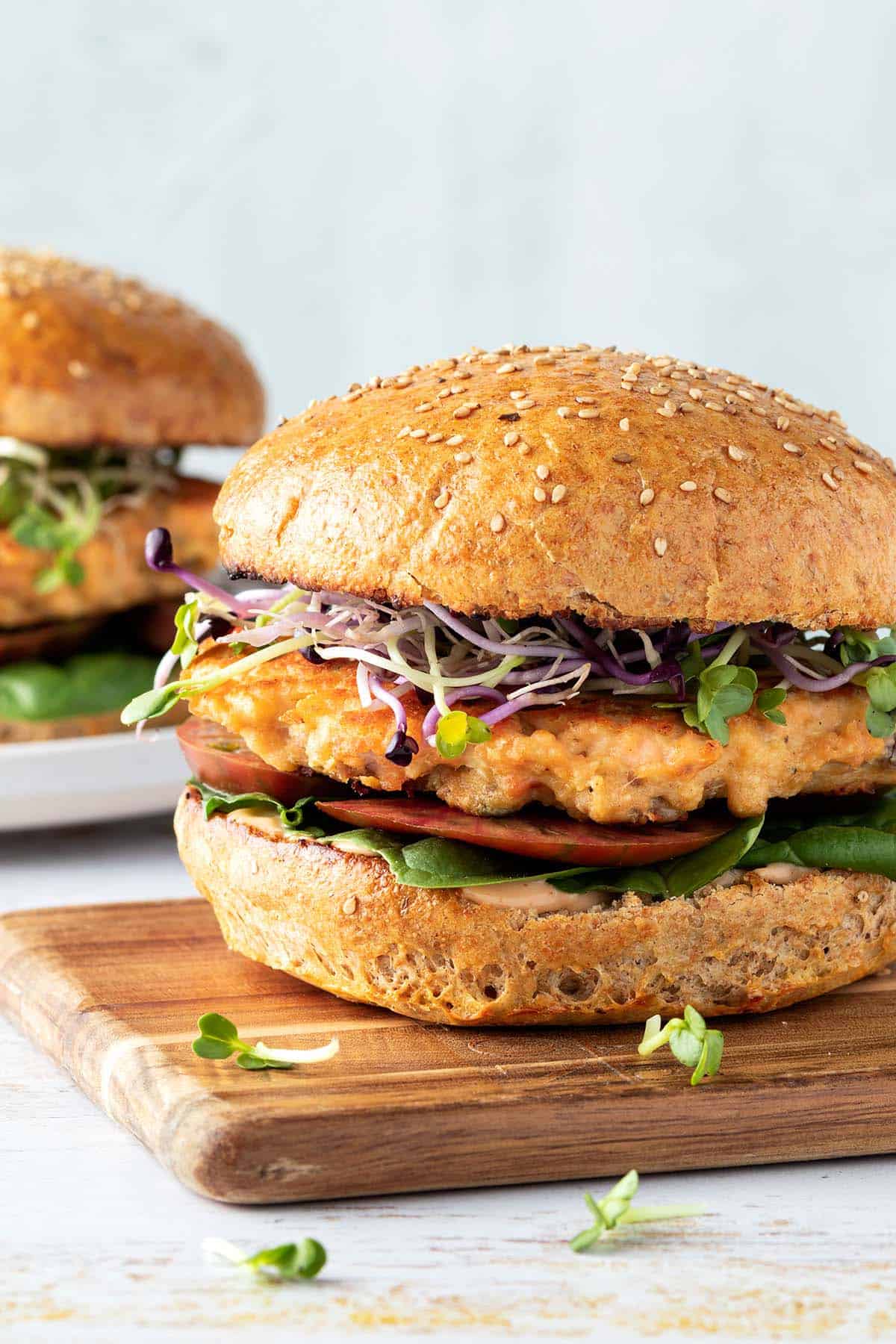 Ein saftiger Lachsburger mit einem goldbraun angebratenen Lachs Patty, frischen Sprossen, Tomaten und Salat auf einem Holzbrett, im Hintergrund ein zweiter Burger.