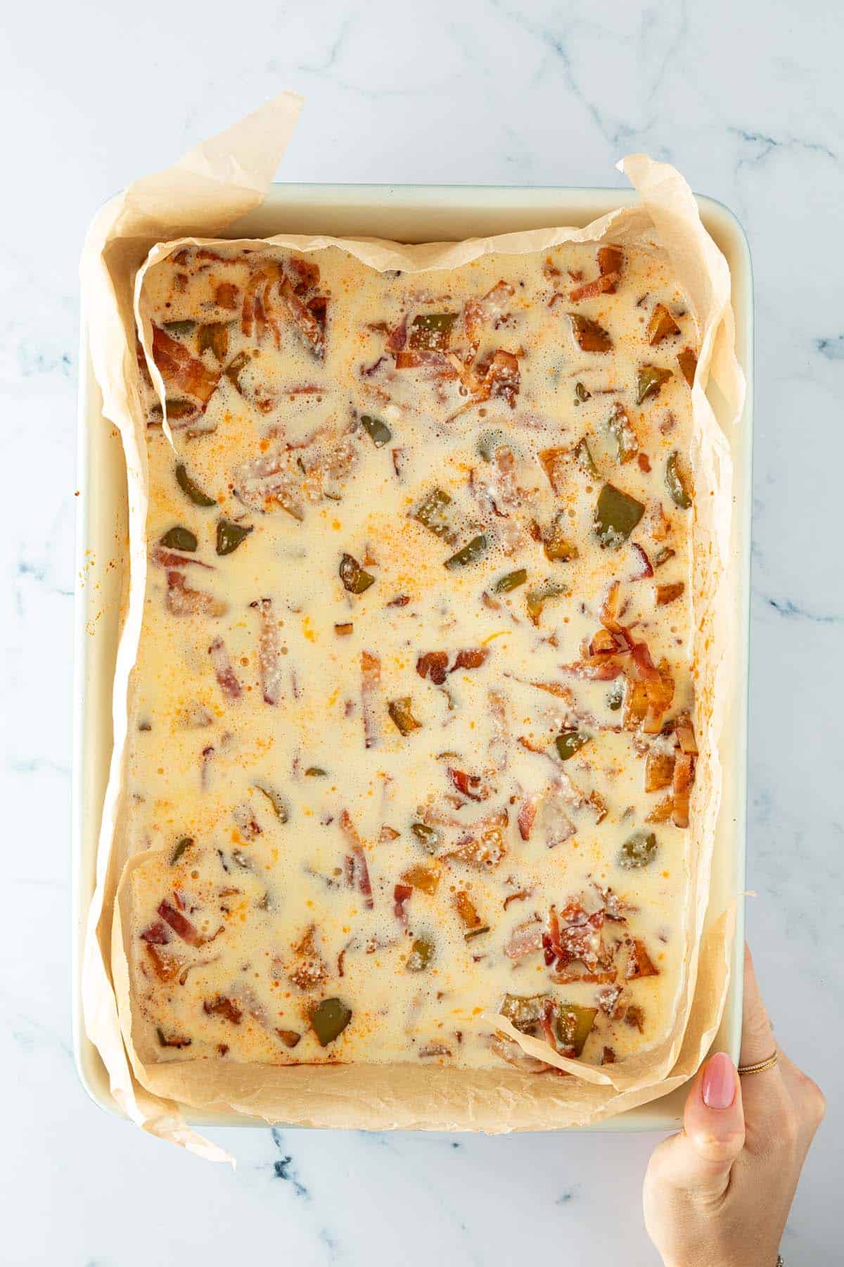 Auflaufform mit Backpapier ausgelegt und gefüllt mit roher Eier-Hüttenkäse-Mischung, Kartoffeln, Paprika und Speckstreifen.