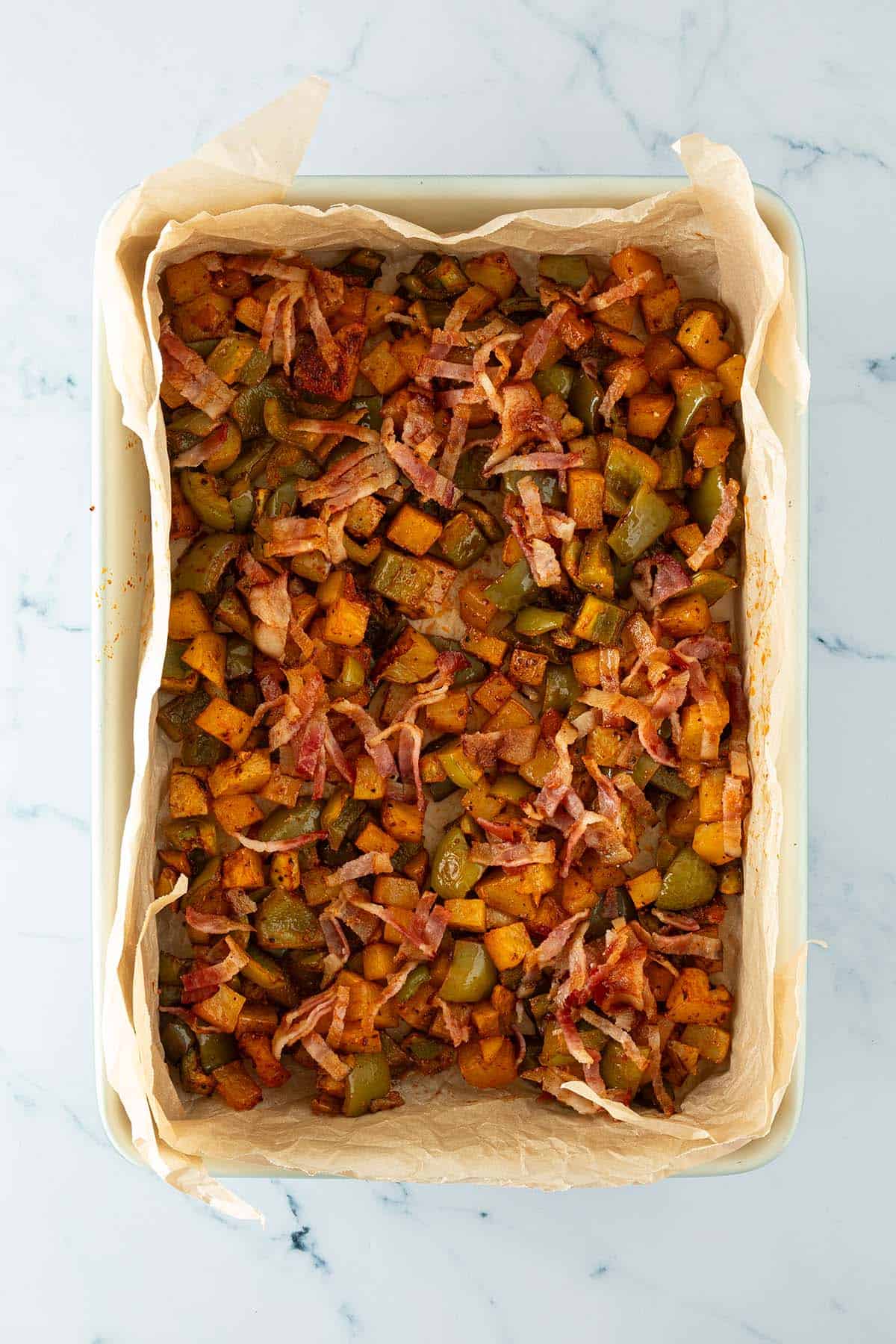 Geröstete Kartoffelwürfel, grüne Paprika und knusprige Speckstreifen in einer mit Backpapier ausgelegten Auflaufform.