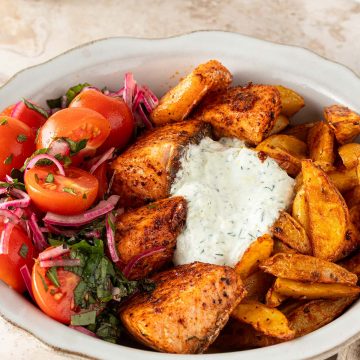 Mediterrane Lachs Bowl mit würzigen Lachswürfeln, knusprigen Kartoffelspalten, Tomatensalat und cremigem Tzatziki auf einem hellen Steinuntergrund.