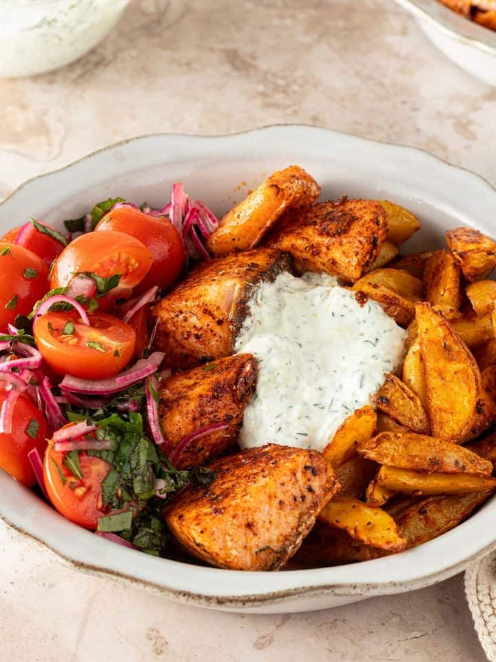 Mediterrane Lachs Bowl mit würzigen Lachswürfeln, knusprigen Kartoffelspalten, Tomatensalat und cremigem Tzatziki auf einem hellen Steinuntergrund.