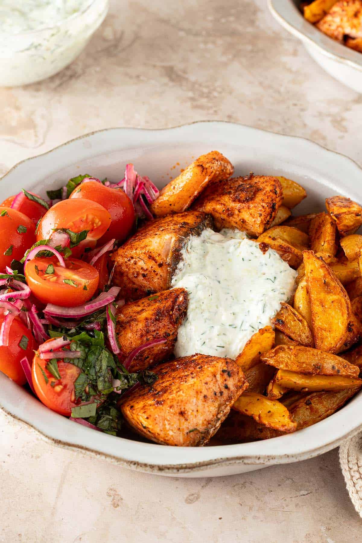 Mediterrane Lachs Bowl mit würzigen Lachswürfeln, knusprigen Kartoffelspalten, Tomatensalat und cremigem Tzatziki auf einem hellen Steinuntergrund.