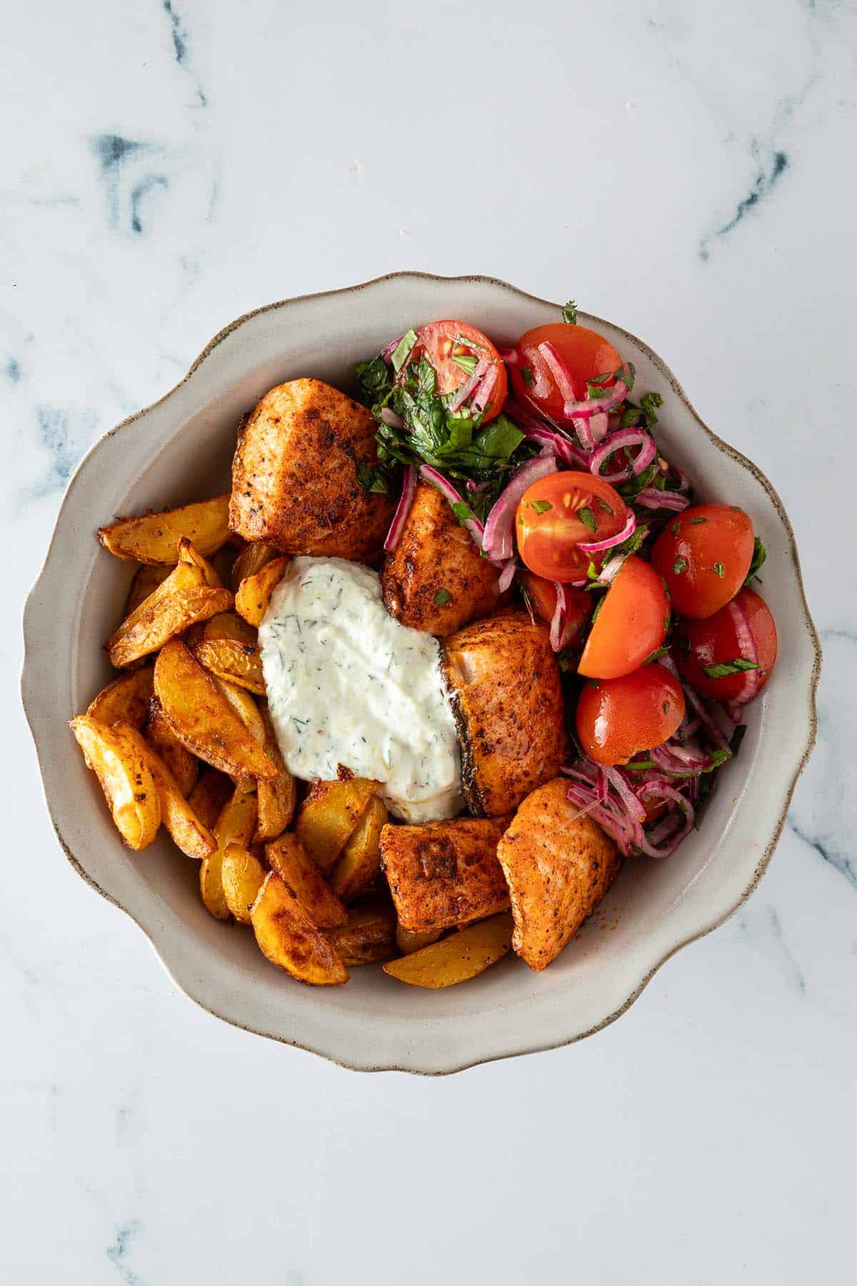Fertig zusammengestellte Bowl aus knusprigen Kartoffelspalten, Lachs-Würfeln, Tomatensalat und Tzatziki.