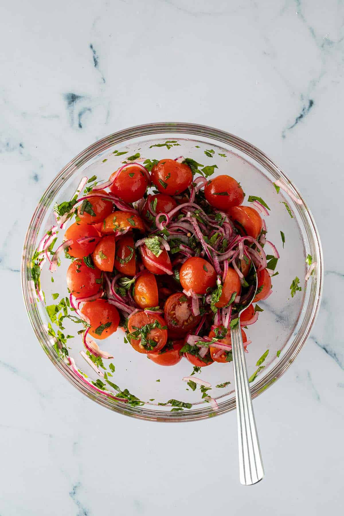 Tomatensalat mit halbierten Cherrytomaten, marinierten roten Zwiebeln und frischen Kräutern in einer Glasschüssel.