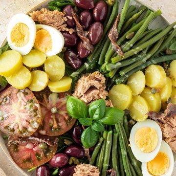 Bunter Nizzasalat serviert auf einem großen Teller mit Bohnen, Kartoffeln, Oliven, Sardellen, Thunfisch, Eiern, Tomaten und Kapern auf einem hellen Untergrund.