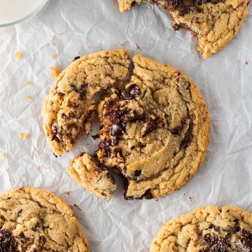 Vier große Chocolate Chip Cookies auf Backpapier, einer angebissen, einer angebrochen mit sichtbar geschmolzener Schokolade, daneben ein Glas Milch.