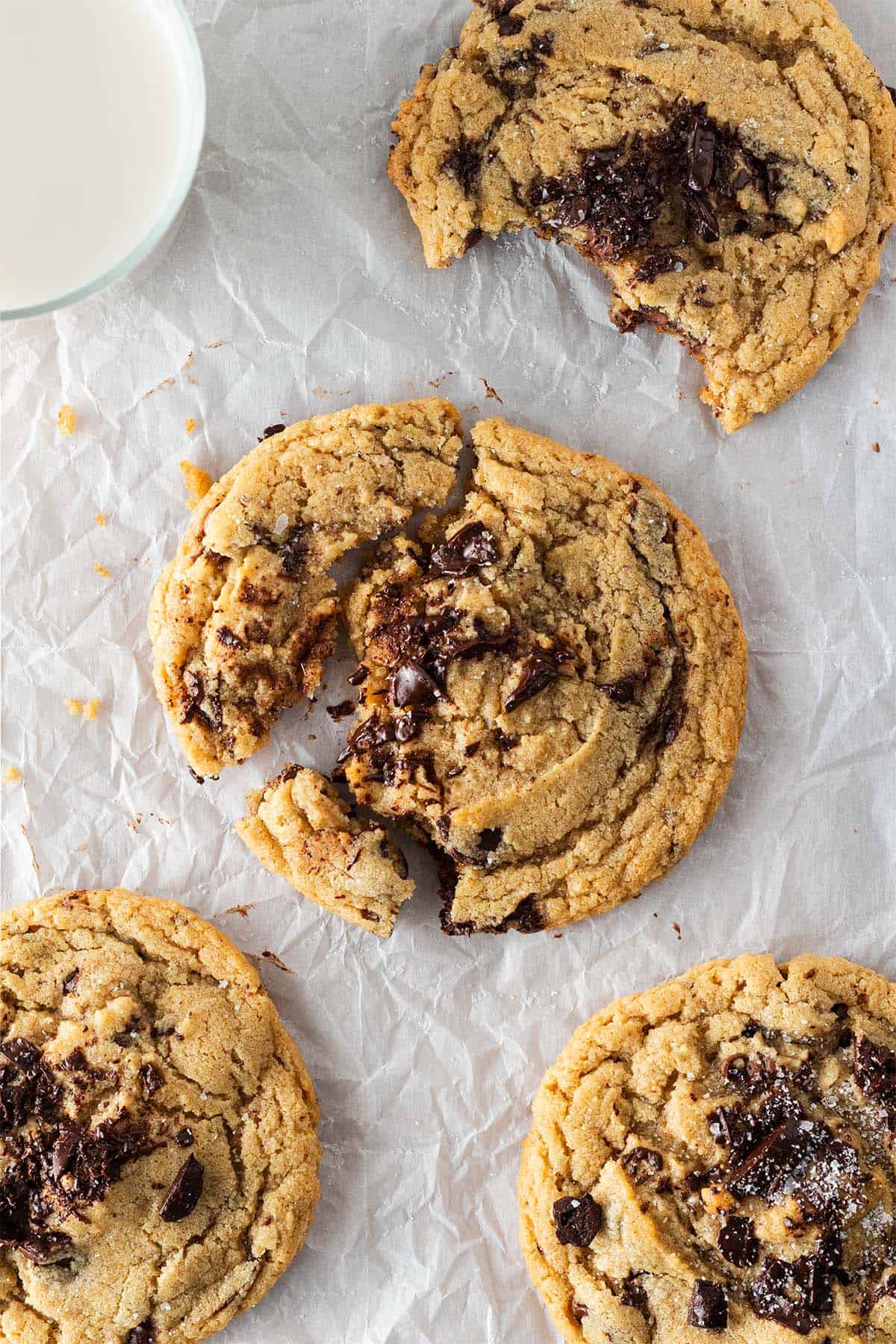 Vier große Chocolate Chip Cookies auf Backpapier, einer angebissen, einer angebrochen mit sichtbar geschmolzener Schokolade, daneben ein Glas Milch.