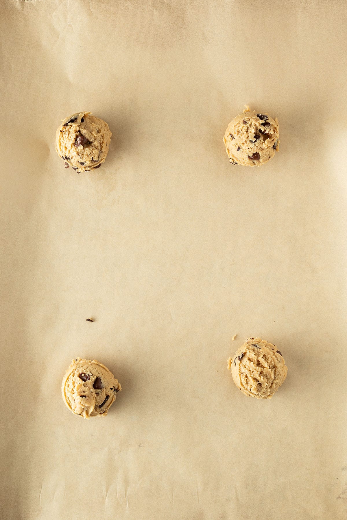 Vier Kugeln Chocolate-Chip-Cookie-Teig gleichmäßig auf Backpapier verteilt, vor dem Backen.