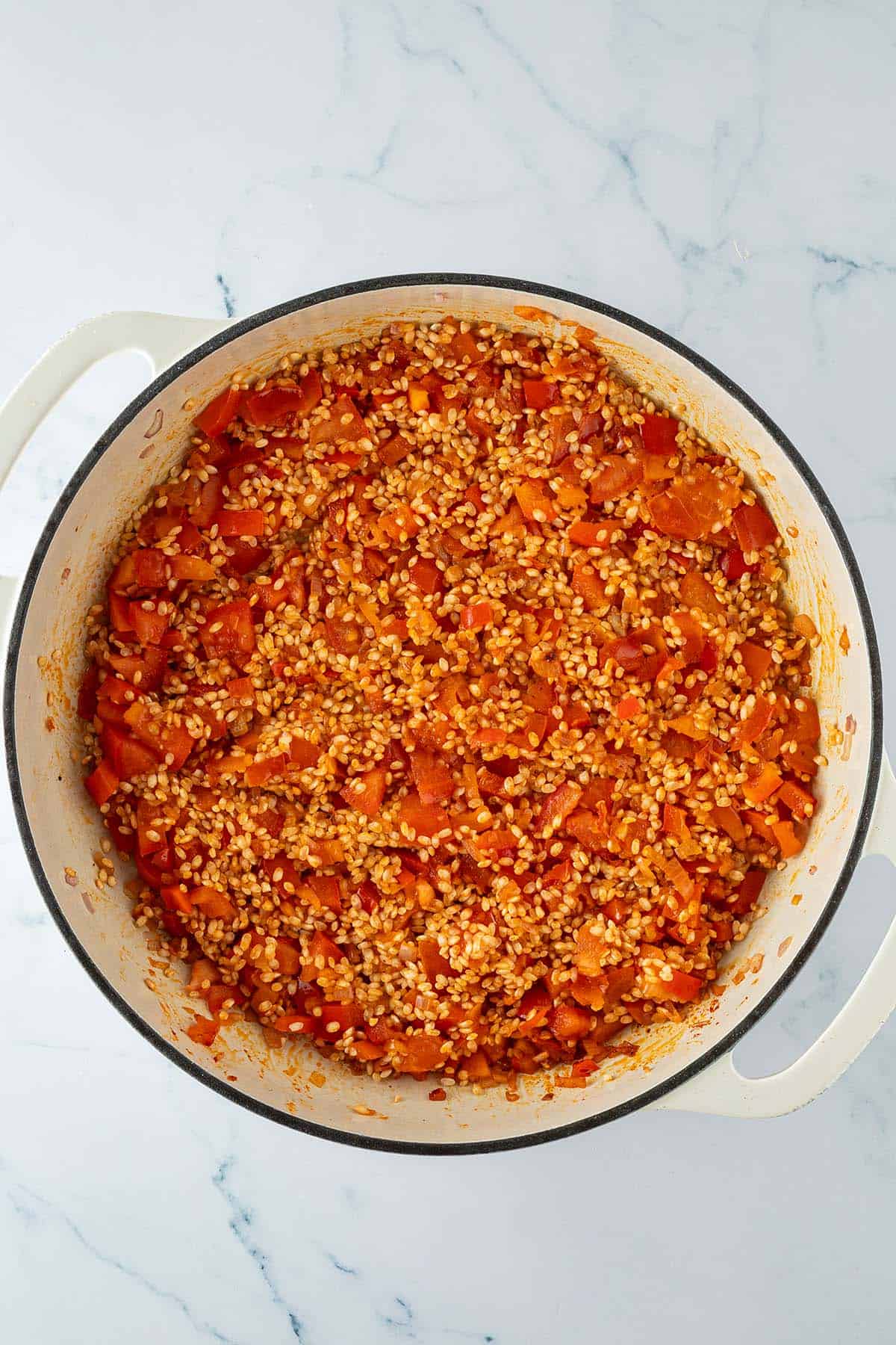 Ungekochter Paella-Reis mit angebratener Paprika, Tomaten und Zwiebeln in einer großen Pfanne.