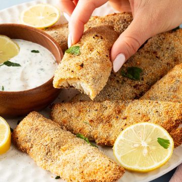 Hand greift nach einem knusprig panierten Fischfilet mit Zitronenscheiben und Dip auf einem Teller.