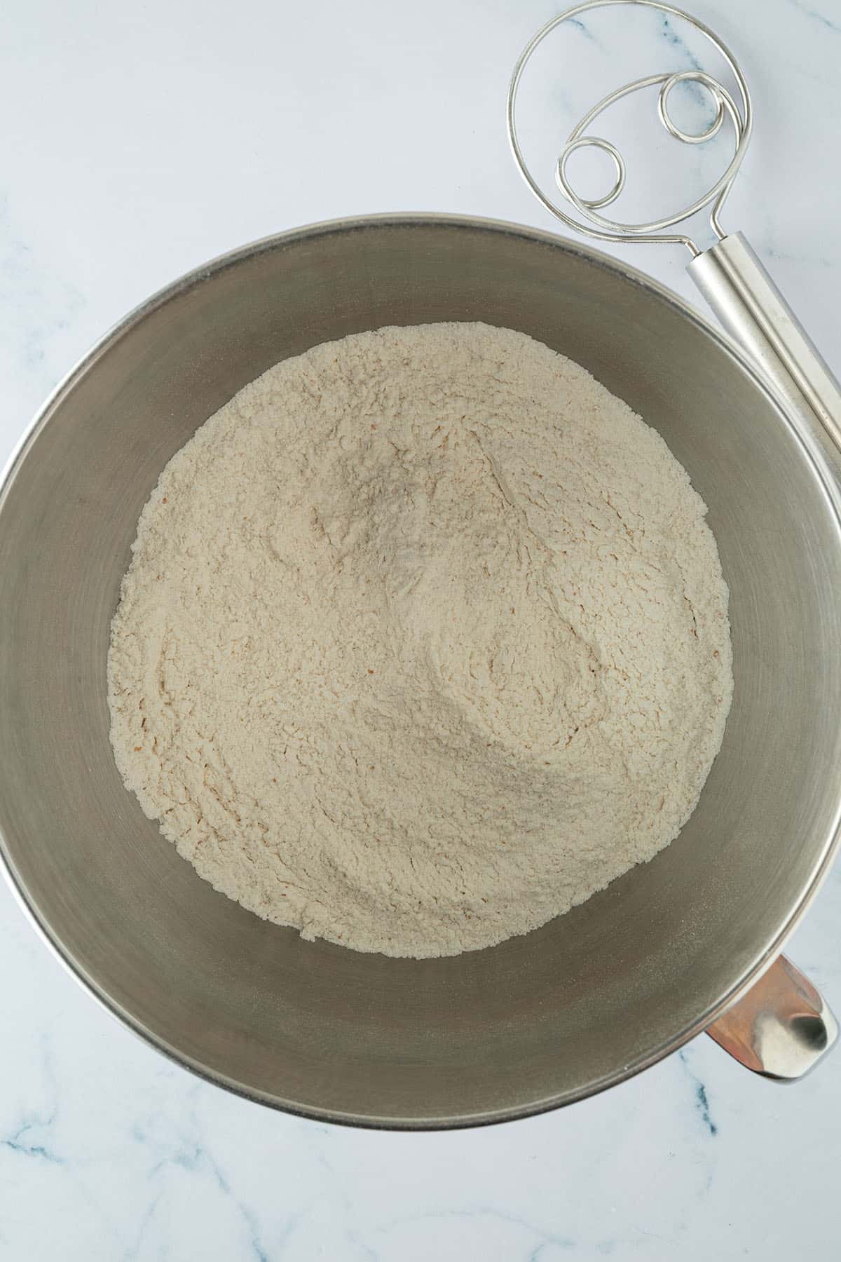 Trockene Zutaten f&uuml;r Pinsa Teig in einer R&uuml;hrsch&uuml;ssel vermischt, daneben liegt ein d&auml;nischer Schneebesen.