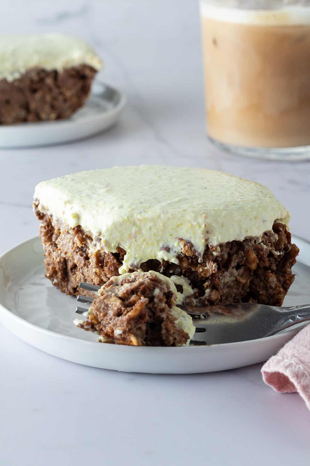 Ein St&uuml;ck Schoko Pistazien Baked Oatmeal auf einem Teller, ein Bissen davon auf einer Gabel mit einem Eiskaffee und einem zweiten St&uuml;ck im Hintergrund.