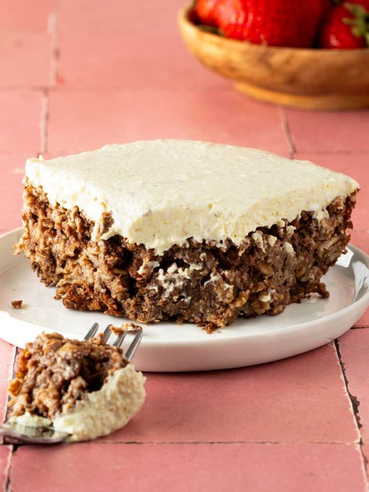 EIn Stück Pistazien Baked Oatmeal auf einem Teller, ein Stück davon auf einer Gabel liegt davor, eine Schüssel Erdbeeren im Hintergrund.