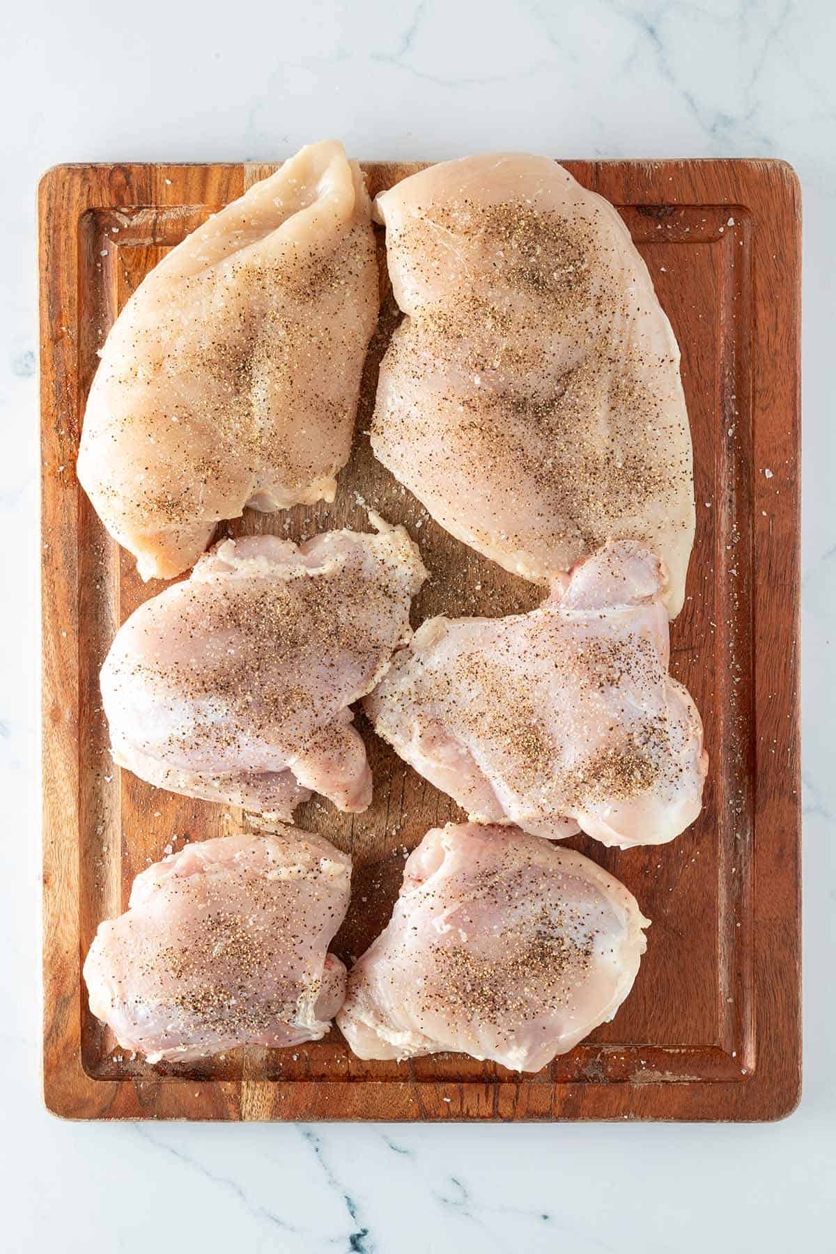 Mit Salz und Pfeffer gew&uuml;rzte, rohe H&auml;hnchenfilets und Oberkeulen auf einem Holzbrett.