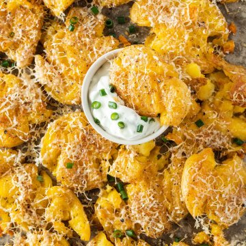 Nahaufnahme von mit Parmesan überbackene Quetschkartoffeln mit einer kleinen Sourcream-Schüssel in der Mitte des Blechs.