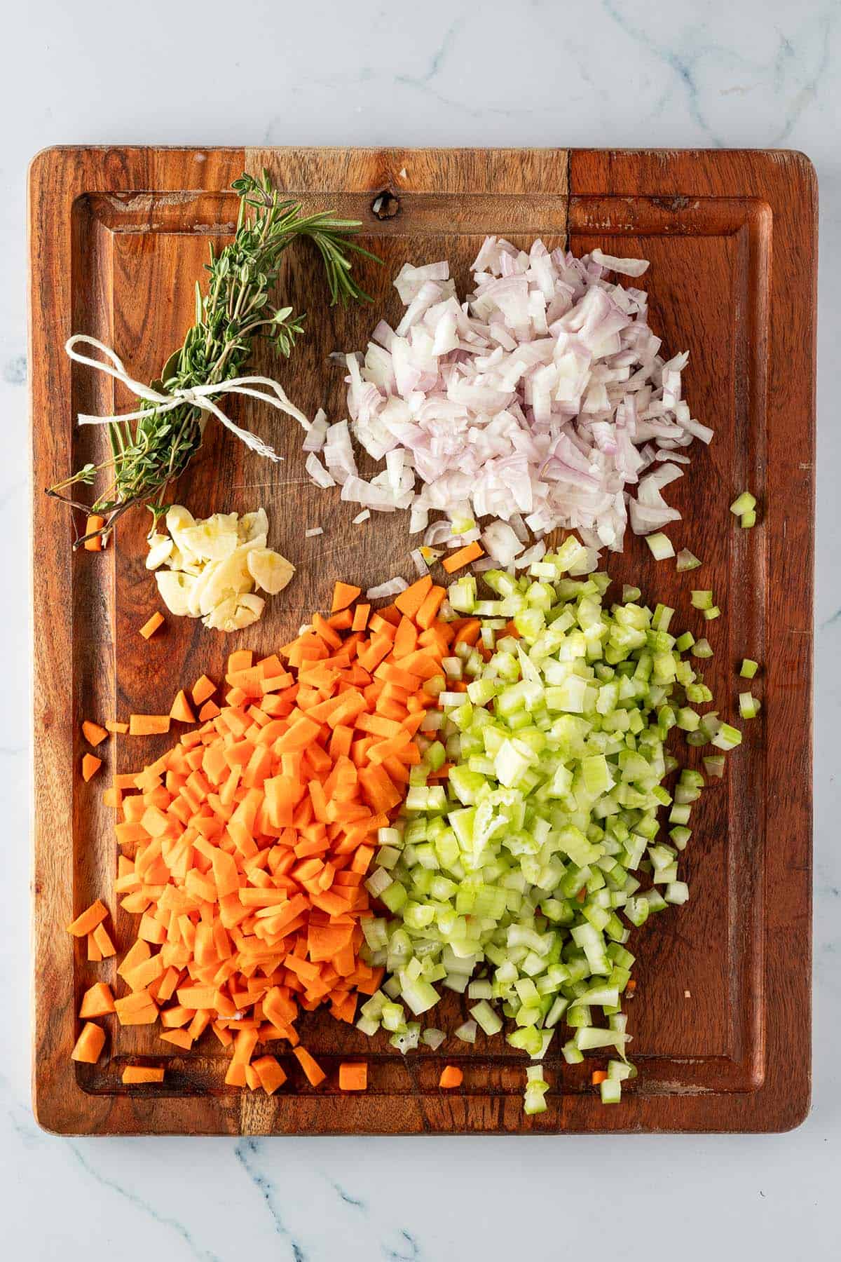 Fein gewürfelte Karotten, Sellerie, Schalotten, in dünne Scheiben geschnittener Knoblauch und zu einem Bündel gebundene Kräuter auf einem Holzbrett.