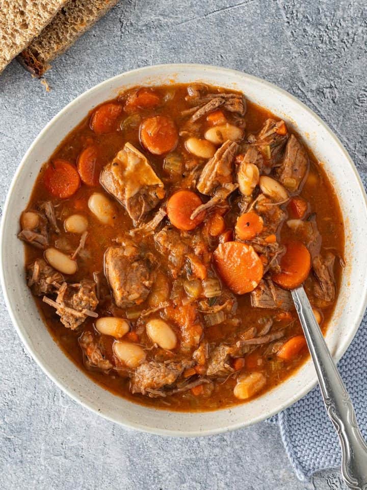 Eine Schüssel Rindfleischeintopf mit Karotten und weißen Bohnen auf einem grauen Untergrund, zwei Scheiben Brot im Bildrand.
