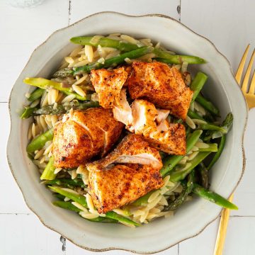 Orzo-Pasta mit gr&uuml;nem Spargel und saftig gebratenem Lachs in einer Sch&uuml;ssel, serviert auf hellem Tisch mit Glas und Gabel.
