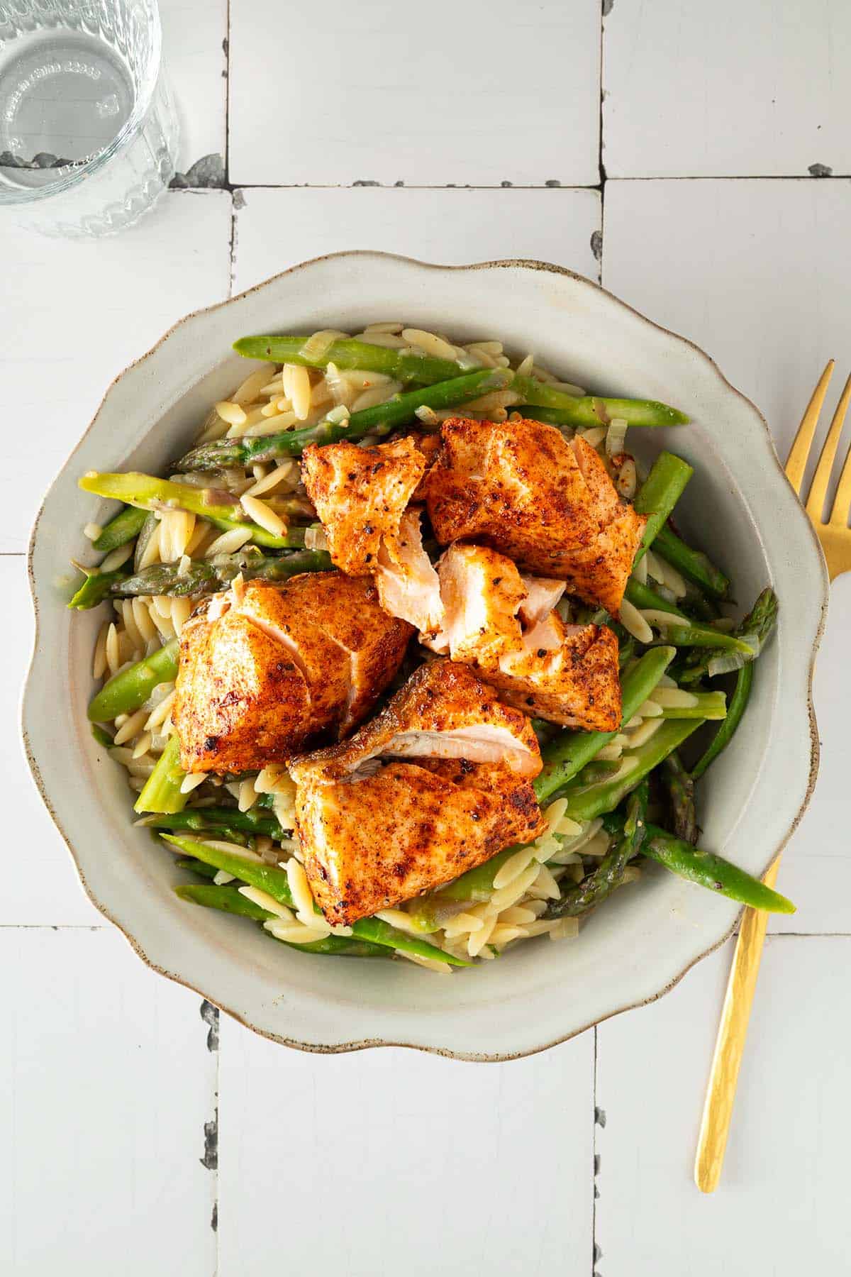 Orzo-Pasta mit gr&uuml;nem Spargel und saftig gebratenem Lachs in einer Sch&uuml;ssel, serviert auf hellem Tisch mit Glas und Gabel.