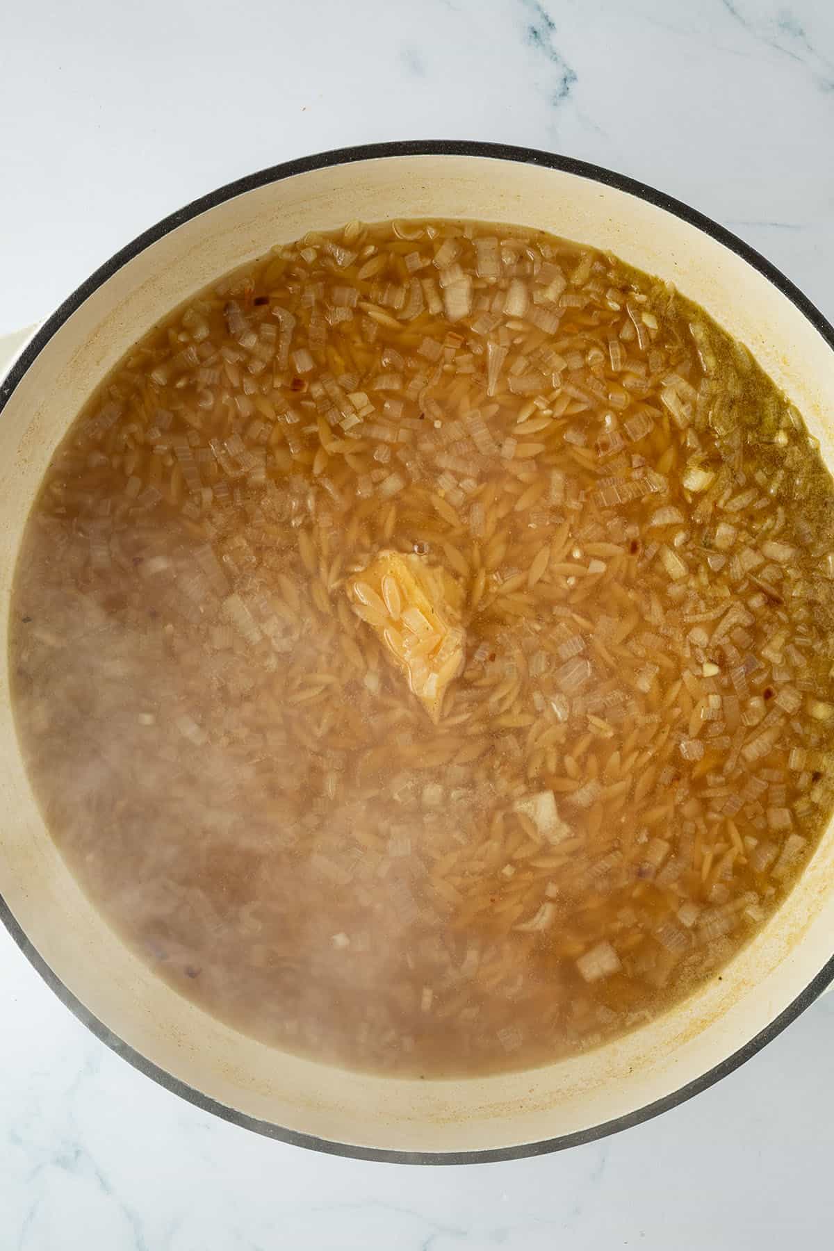 Orzo-Nudeln mit Zwiebeln und einem St&uuml;ck Parmesanrinde in Br&uuml;he k&ouml;cheln in einer Pfanne