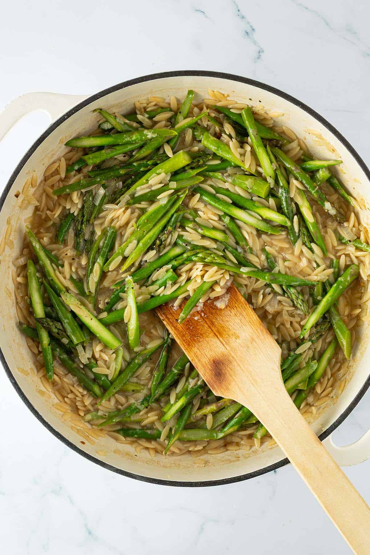 Cremige Orzo mit gr&uuml;nem Spargel in der Pfanne, wird mit einem Holzl&ouml;ffel ger&uuml;hrt.