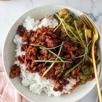 Bowl mit wei&szlig;em Kokosreis, knusprig gebratenem Sticky Tempeh und w&uuml;rzigem Gurkensalat, garniert mit Fr&uuml;hlingszwiebeln und serviert mit einer Gabel.