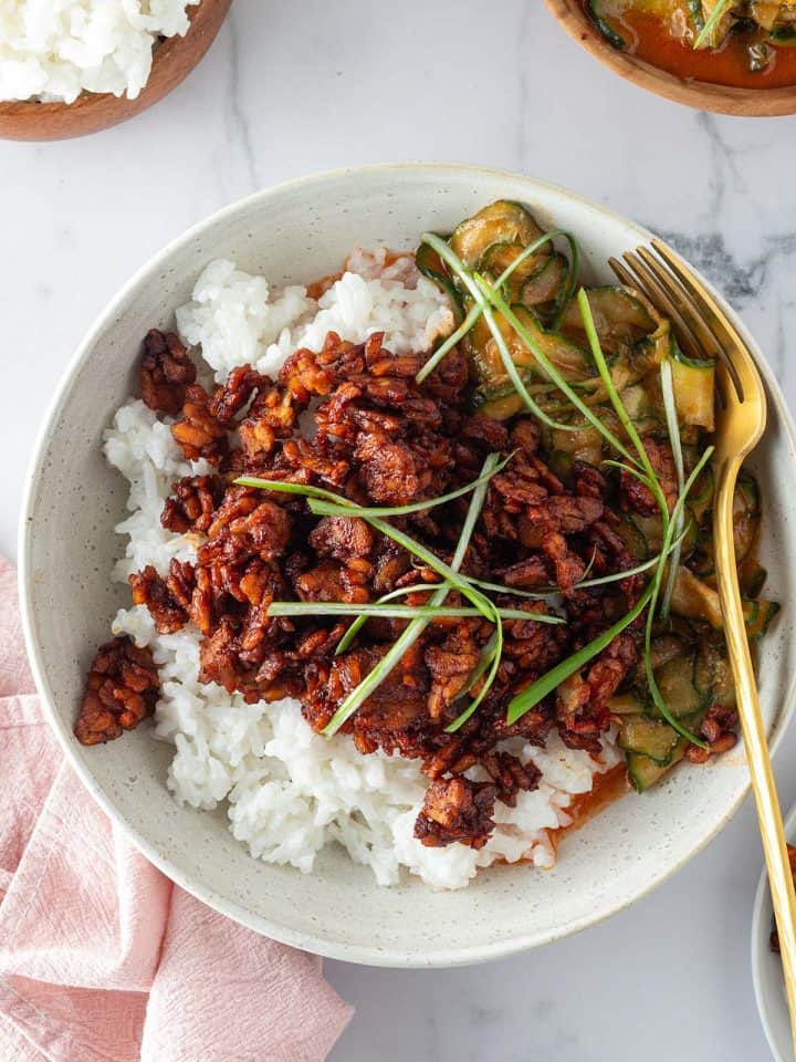 Bowl mit weißem Kokosreis, knusprig gebratenem Sticky Tempeh und würzigem Gurkensalat, garniert mit Frühlingszwiebeln und serviert mit einer Gabel.