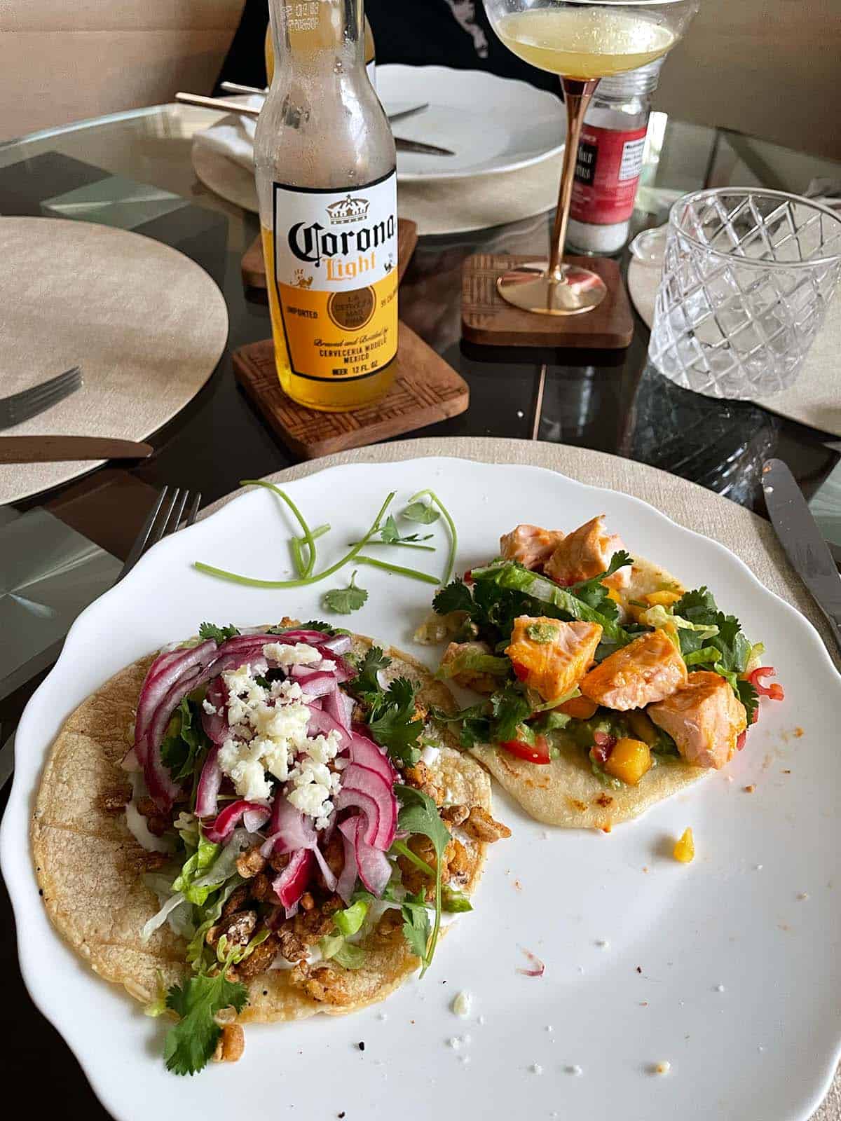 Zwei unterschiedlich belegte Tacos auf einem Esstisch, Margaritas und eine Corona Flasche sowie mehrere Teller im Bildhintergrund.