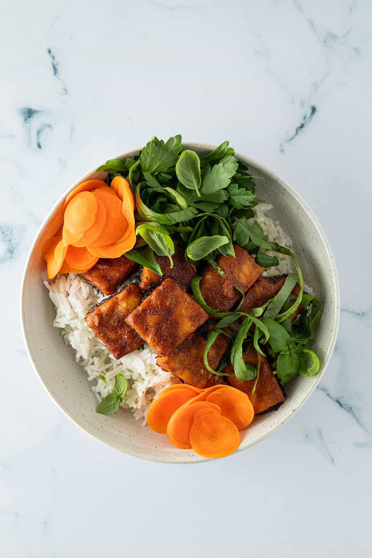 Reis, frische Kräuter, eingelegte Karotten und knusprig gebackener Tempeh in einer Bowl angerichtet.
