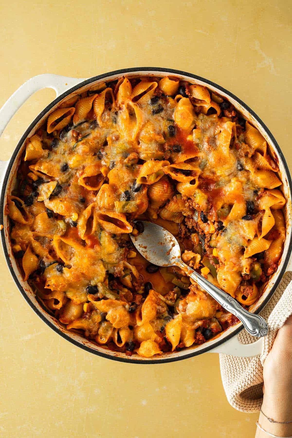 Auflaufform mit überbackenem Tex-Mex Nudelauflauf aus Conchiglie, Hackfleisch, schwarzen Bohnen und geschmolzenem Käse, ein Löffel hebt eine Portion heraus.