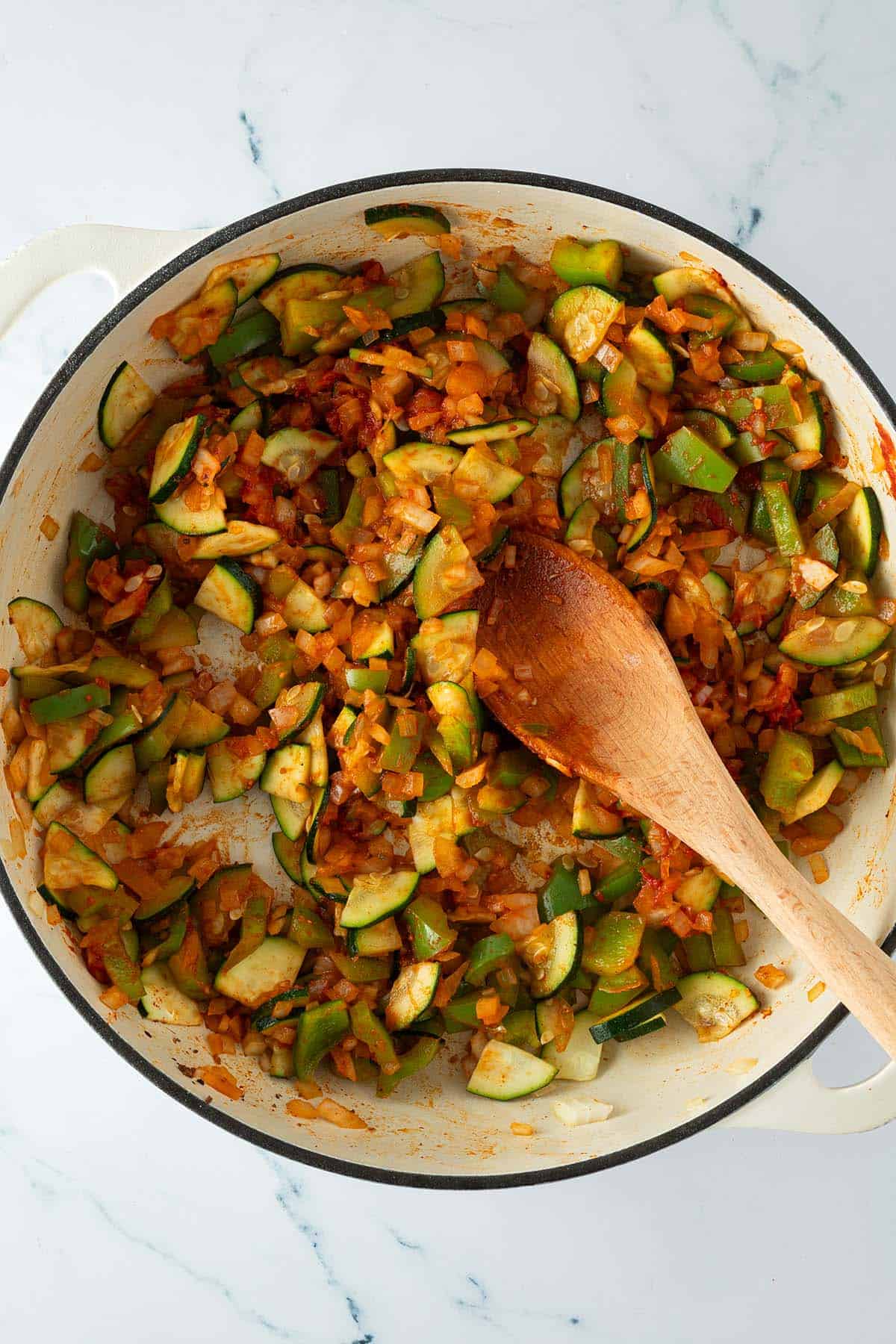 Pfanne mit angebratenem Gemüse aus Zucchini, Paprika, Zwiebeln, Gewürzen und Tomatenmark das mit einem Holzlöffel vermischt wird.