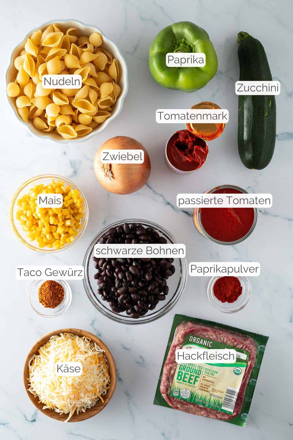 Alle Zutaten mit jeweiligen Textbeschriftungen, die für den Tex Mex Nudelauflauf mit Hackfleisch benötigt werden.