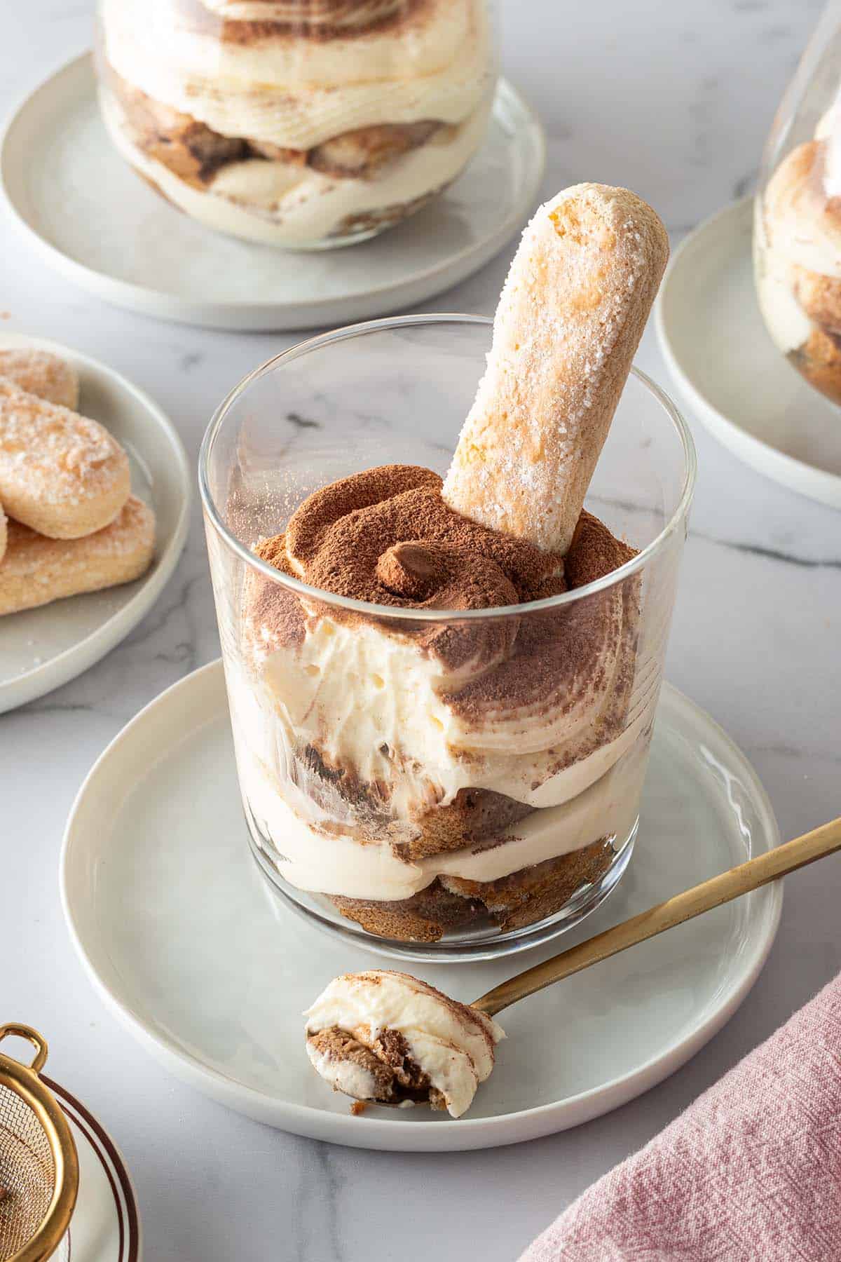Tiramisu im Glas geschichtet mit Löffelbiskuit, im Hintergrund mehr Gläser mit Tiramisu und Löffelbiskuits.
