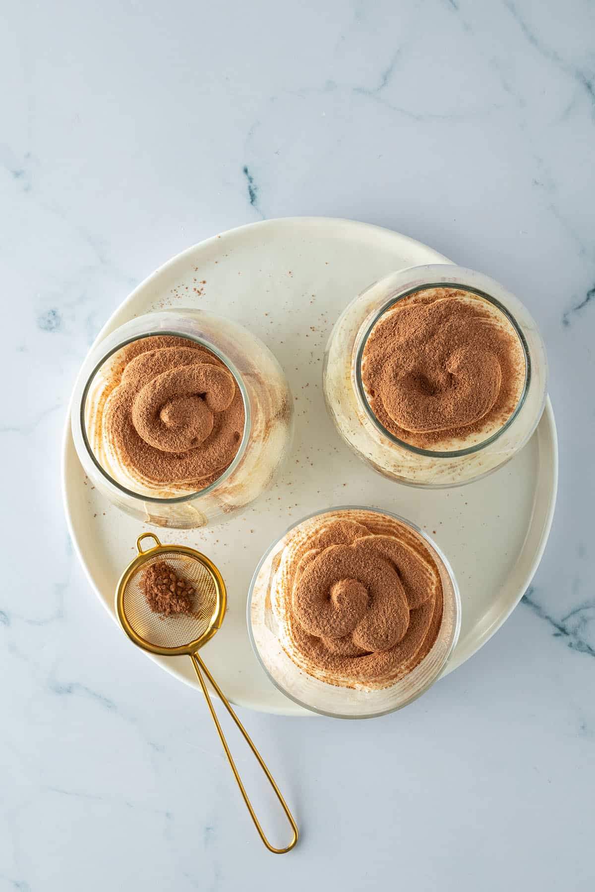 Drei Gläser Tiramisu von oben, bestäubt mit Kakao, daneben ein kleines Sieb.