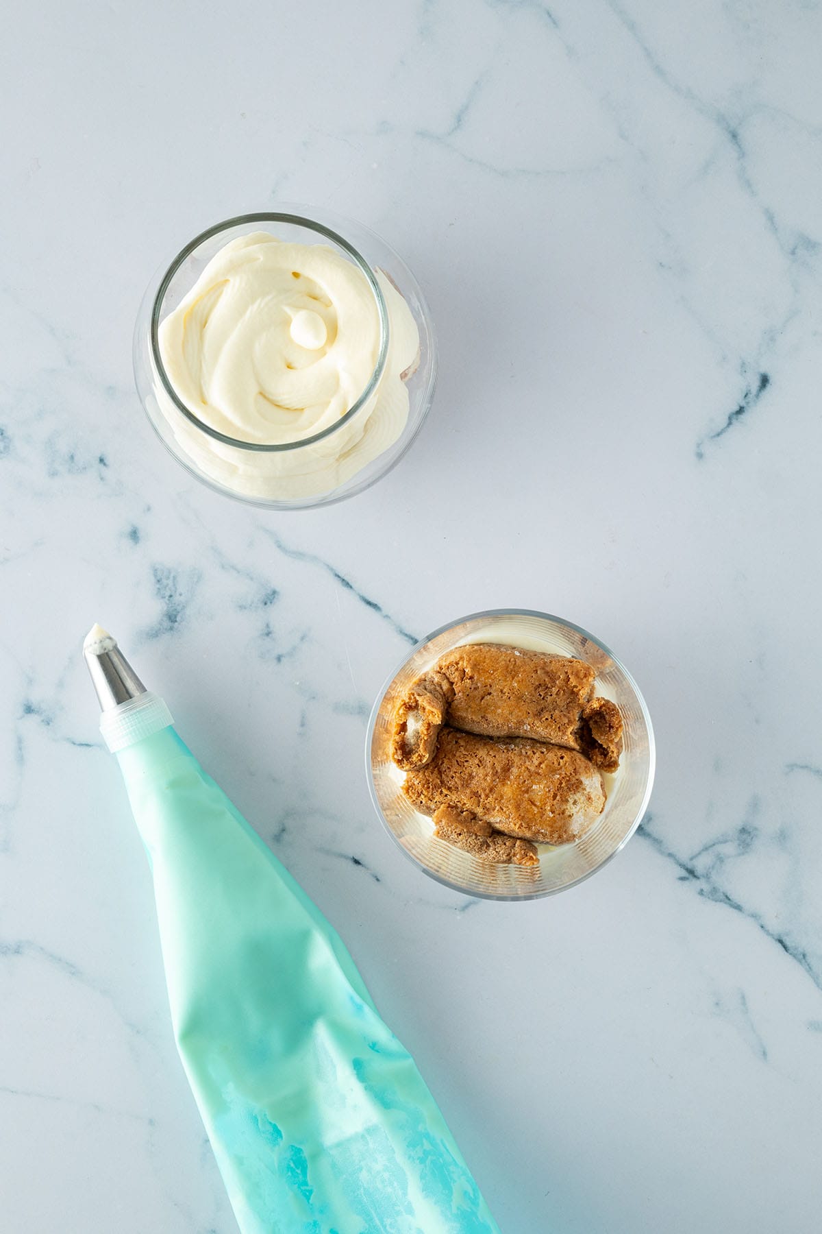 Ein Glas mit getränkten Löffelbiskuits und ein Glas mit Creme, daneben ein gefüllter Spritzbeutel.