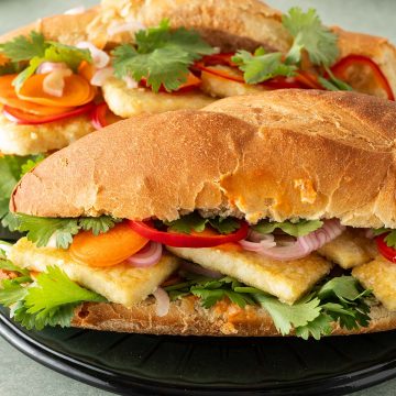 Zwei Tofu Banh Mi Sandwiches mit eingelegtem Gemüse und frischem Koriander auf einem Teller, Koriander und ein Glas im Hintergrund.