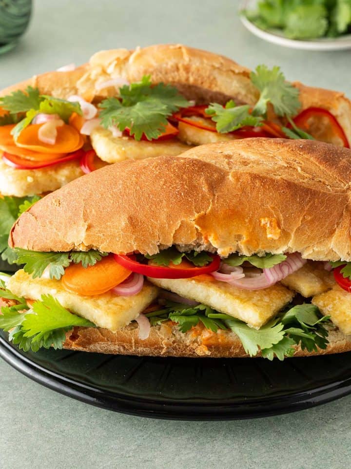 Zwei Tofu Banh Mi Sandwiches mit eingelegtem Gemüse und frischem Koriander auf einem Teller, Koriander und ein Glas im Hintergrund.
