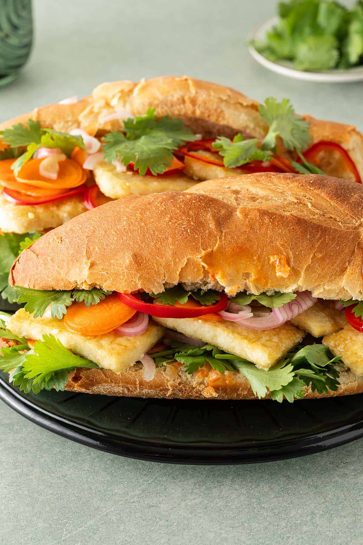 Zwei Tofu Banh Mi Sandwiches mit eingelegtem Gemüse und frischem Koriander auf einem Teller, Koriander und ein Glas im Hintergrund.