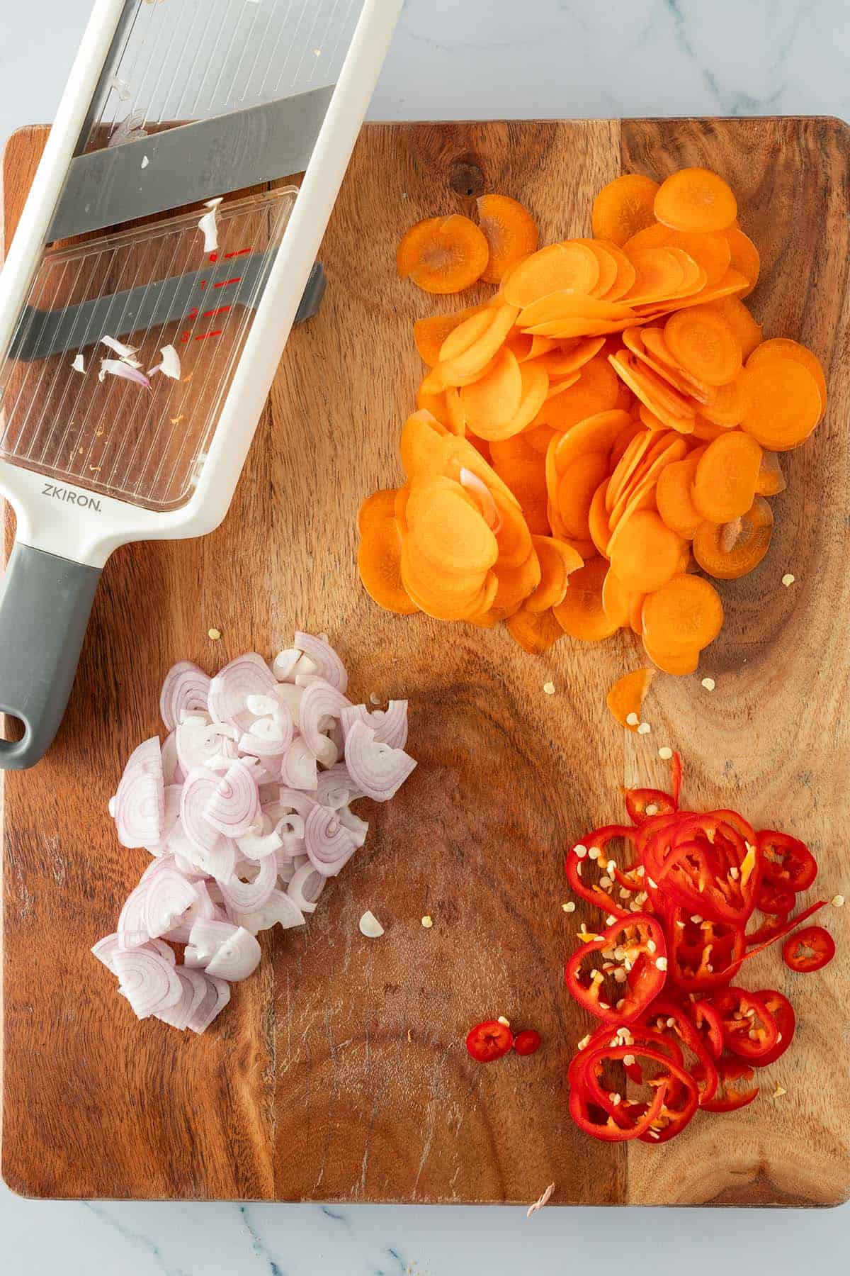 In dünne Scheibchen geschnittene Karotten, Schalotten und rote Chili auf Holzbrett mit einer Gemüsehobel daneben.