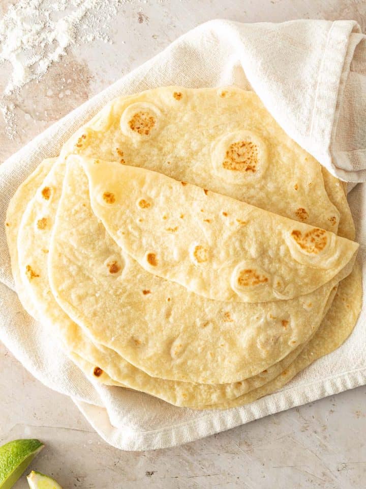 Ein Stapel selbstgemachte Weizen Tortillas auf einem hellen Tuch, mit Mehl und Limetten im Bildrand.