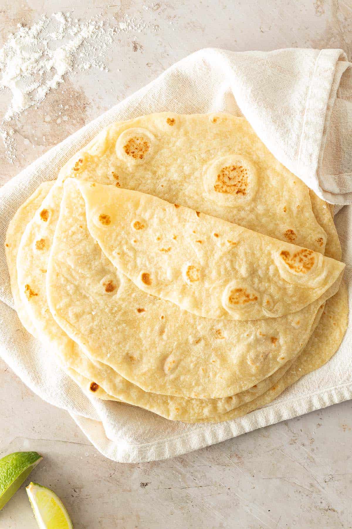 Ein Stapel selbstgemachte Weizen Tortillas auf einem hellen Tuch, mit Mehl und Limetten im Bildrand.